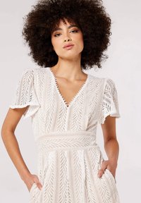 White, lace dress with a V-neck, short ruffled sleeves, intricate patterns, and inset pockets. Textured design enhances visual appeal.