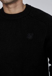 Črn pleten pulover z žametno teksturo, okroglo ovratnico in majhnim tonom usklajenim logotipom na prsih. Visokokakovosten material, sproščen dizajn.
