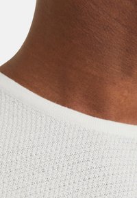 Weißer, strukturierten Stoff mit einem gestrickten Design, ergänzt durch einen glatten Halsausschnitt mit einem kontrastierenden, hellen Saum.