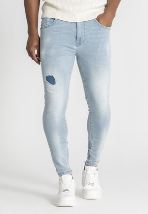 Jeans skinny bleu clair avec un détail déchiré au niveau du genou, fabriqués en denim, présentant une texture lisse et un léger dégradé. Associés à des baskets blanches.