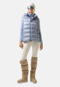 Helleblaue gesteppte Weste mit Kapuze, kombiniert mit einem passenden langärmligen Oberteil und cremfarbigen Leggings. Tanfarbene, plüschige Stiefel und eine gestrickte weiße Mütze.