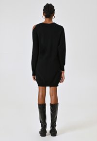 Donna con i capelli intrecciati che indossa un vestito maglione nero con spalle scoperte e stivali alti in pelle nera, in piedi di spalle.