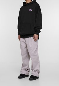 Musta huppari, jossa on etutasku ja vaaleanpunainen logo, yhdistetty vaaleanharmaisiin leveälahkeisiin housuihin ja mustiin tennareihin valkoisella taustalla.