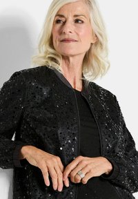 Veste bomber noire avec des sequins, fermeture zippée et poignets côtelés. Fini texturé avec des accents brillants. Association avec un haut noir visible en dessous.