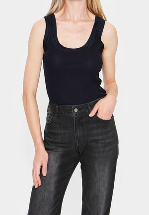 Frau trägt ein schwarzes ärmelloses Rippoberteil und hoch taillierte ausgewaschene schwarze Jeans, steht mit einer Hand in der Tasche vor weißem Hintergrund.