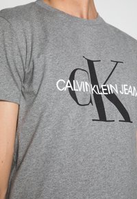 Grå bomullströja med korta ärmar, med en stor svart "CK"-logotyp och vit text "CALVIN KLEIN JEANS" över bröstet.