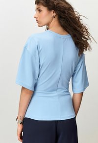Lyseblå kortærmet top med rund hals, figursyet talje og detalje med søm bagpå. Blødt, glat stof med afslappet silhuet.