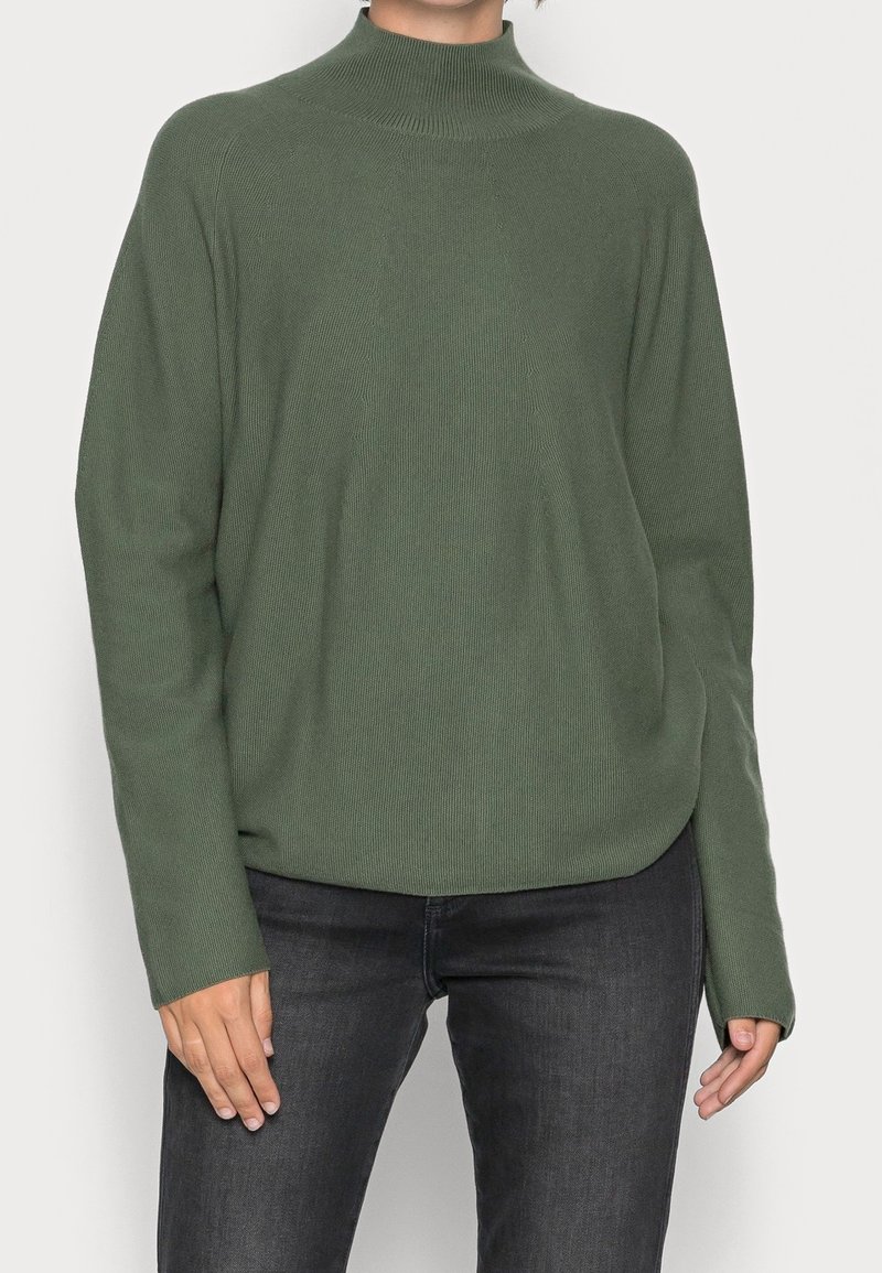 Personne portant un pull vert olive côtelé à manches longues avec un col montant et un jean gris foncé, debout devant un fond clair uni.