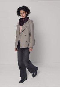 Grijs dubbelgeknoopt colbert met zwarte knopen, gecombineerd met een donkere sjaal, licht topje en donkere uitlopende jeans. Zwarte schoenen maken de outfit compleet.