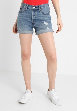 Personne portant un short en denim bleu retroussé avec de petites déchirures, un haut rouge et des baskets blanches se tenant sur un sol blanc.