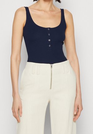 Vrouw die een marineblauwe mouwloze geribde top met knopen aan de voorkant draagt en witte hoge taillebroek met een ritssluiting aan de voorkant.