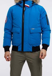 Blå vinterjacka med pälsmössa, svart dragkedja, framfickor med lock och logotypmärke på vänster ärm, bärs med olivgröna cargobyxor.