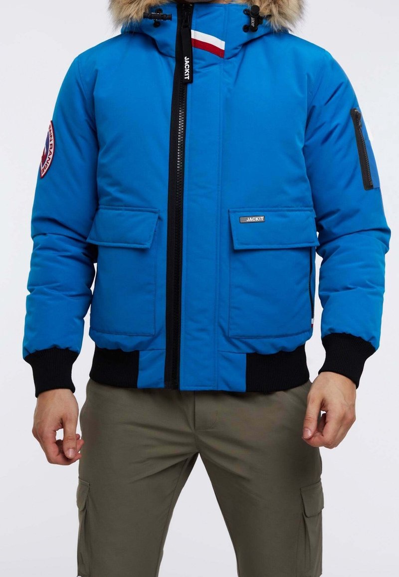 Blå vinterjacka med pälsmössa, svart dragkedja, framfickor med lock och logotypmärke på vänster ärm, bärs med olivgröna cargobyxor.