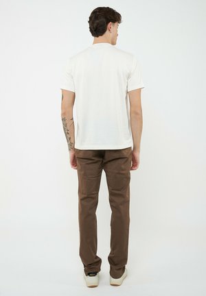 Hombre con tatuaje en el brazo izquierdo, vestido con camisa blanca de manga corta, pantalones marrones y zapatillas blancas, de pie y de espaldas sobre un fondo liso.