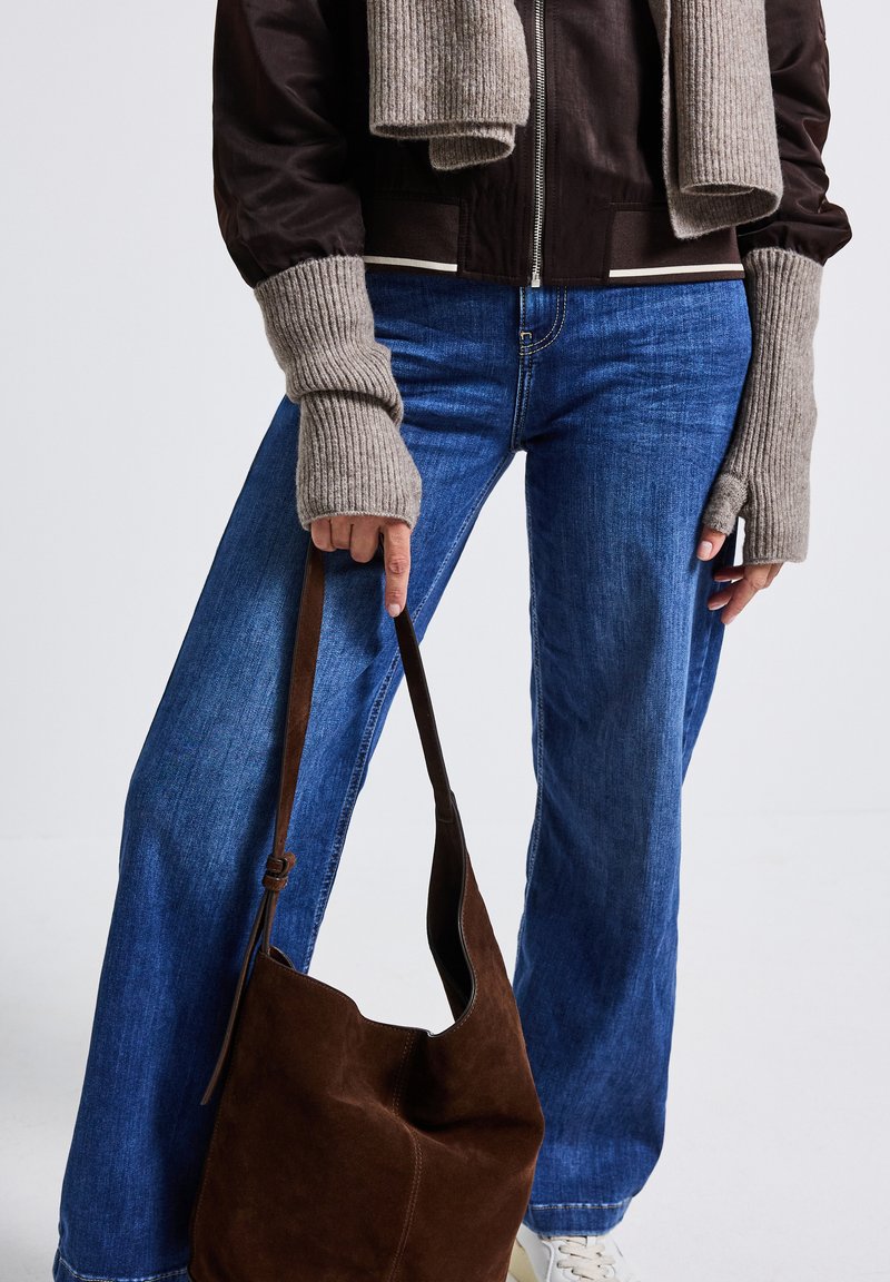 Person med blå jeans, brun jakke, lange beige strikkede fingerløse handsker og tørklæde, der holder en brun ruskinds håndtaske i stropperne.