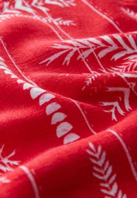 Roter Stoff mit weißen botanischen Mustern, versehen mit Blattmotiven und geschwungenen Designs. Strukturierte Oberfläche, die seine leichte, gewebte Qualität zeigt.