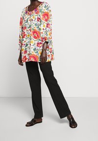 Květinová tunika v živých barvách s lehkým, volným designem, 3/4 rukávy a kulatým výstřihem, kombinovaná s černými kalhotami a sandáli.