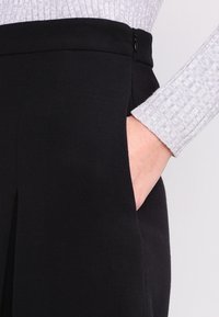 Šedý top s dlouhým rukávem se setkává s černou sukní, přičemž ruka je položená v kapse sukně, což vytváří moderní a stylový detail oblečení.