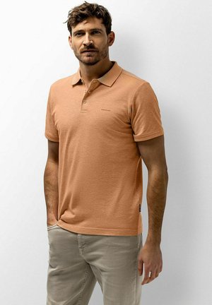 Man met kort bruin haar en baard, die een lichtbruin poloshirt en beige broek draagt, staande tegen een effen witte achtergrond.