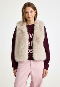 Faux fur vest in lichtbeige met een zachte, pluizige textuur; gedragen over een bordeauxrode trui met lichtroze tekst. Gecombineerd met lichtroze broeken.