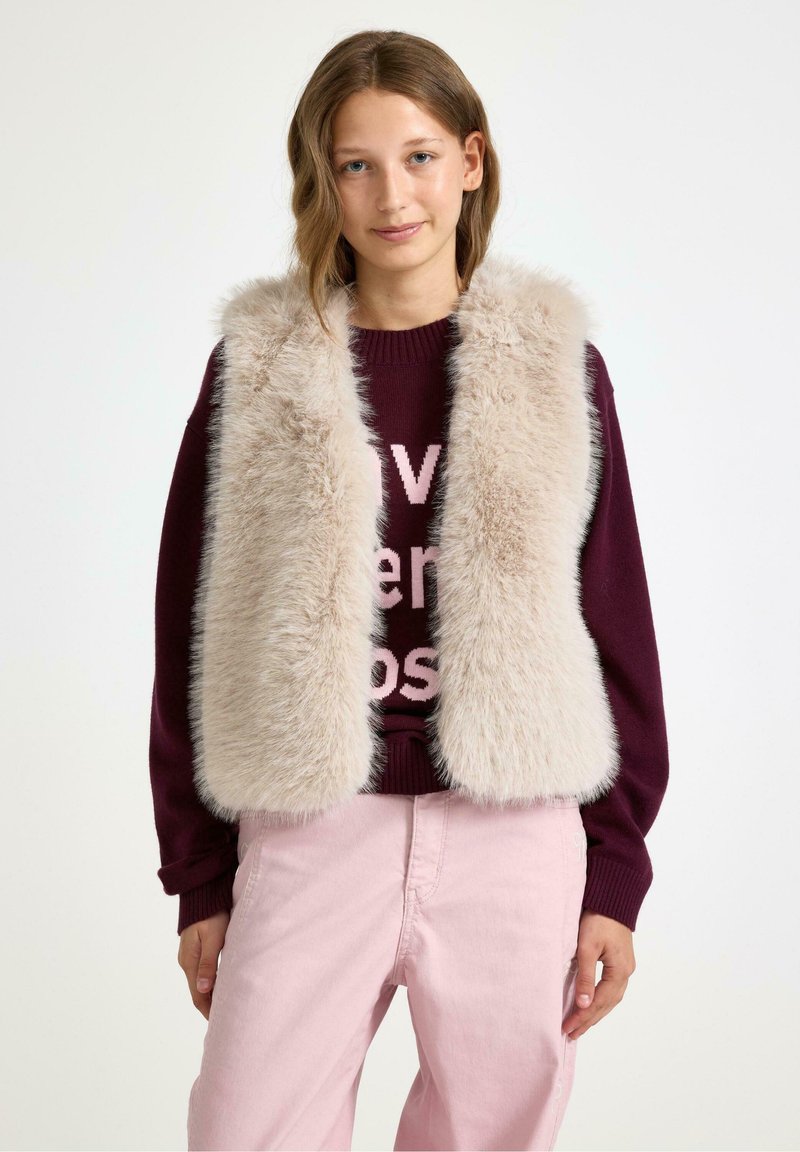 Faux fur vest in lichtbeige met een zachte, pluizige textuur; gedragen over een bordeauxrode trui met lichtroze tekst. Gecombineerd met lichtroze broeken.