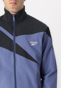 Reebok jakke med en sort og lys lilla farveskala, lynlås på foran, høj krave og vinklet farveblokdesign.
