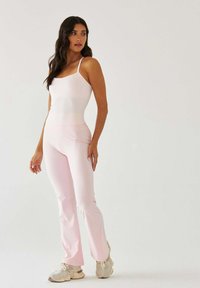 Jasnoróżowy top na ramiączkach z regulowanymi cienkimi paskami, zestawiony z dopasowanymi, rozszerzanymi spodniami. Tkanina wydaje się gładka i elastyczna.