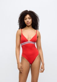 Κόκκινο ολόσωμο μαγιό με λεπτές τιράντες, βαθύ ντεκολτέ και οριζόντια λευκή και μαύρη ριγέ ταινία στη μέση. Λείο ύφασμα.