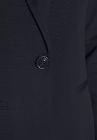 Blazer marine en tissu structuré, fermeture à un bouton noir, et design épuré avec des détails de poche subtils.