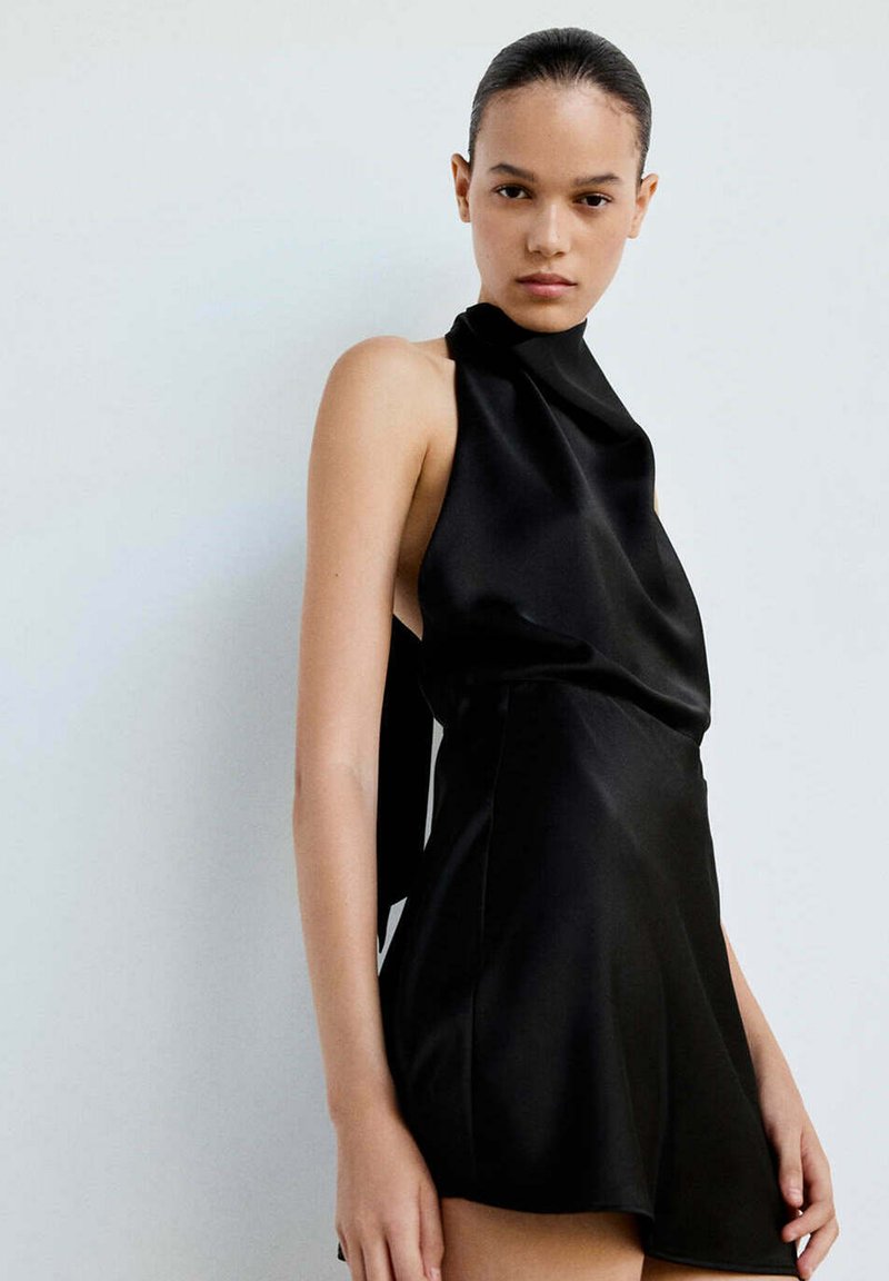 Femme aux cheveux plaqués en arrière portant une robe en satin noire sans manches avec un col montant sur un fond clair uni.