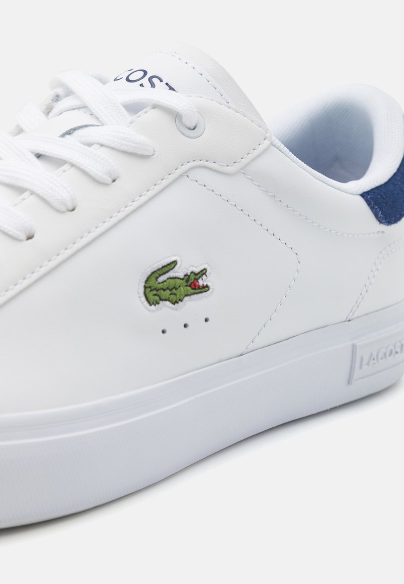 Witte leren sneaker met blauwe suède accenten, voorzien van een groene geborduurde krokodillenlogo en witte veters. Platte rubberen zool.