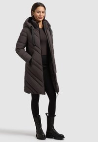 Donna che indossa un lungo cappotto imbottito scuro con cappuccio, leggings neri e stivali neri fino a metà polpaccio, in piedi contro uno sfondo semplice.