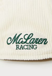 Biely kordurojový látkový štítok s vyšitým logom "McLaren Racing" v zelenej kurzívnej farbe, na bielom oválnom pozadí.