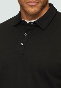 Polo noir en tissu texturé avec col, comportant trois boutons et un design légèrement côtelé.