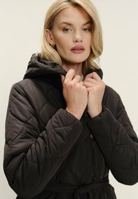 Prešívaná tmavohnedá bunda s kapucňou a viazaním v páse, s textúrovaným vzorom a čiernymi gombíkmi. Rukávy a kapucňa sú mäkké a polstrované.