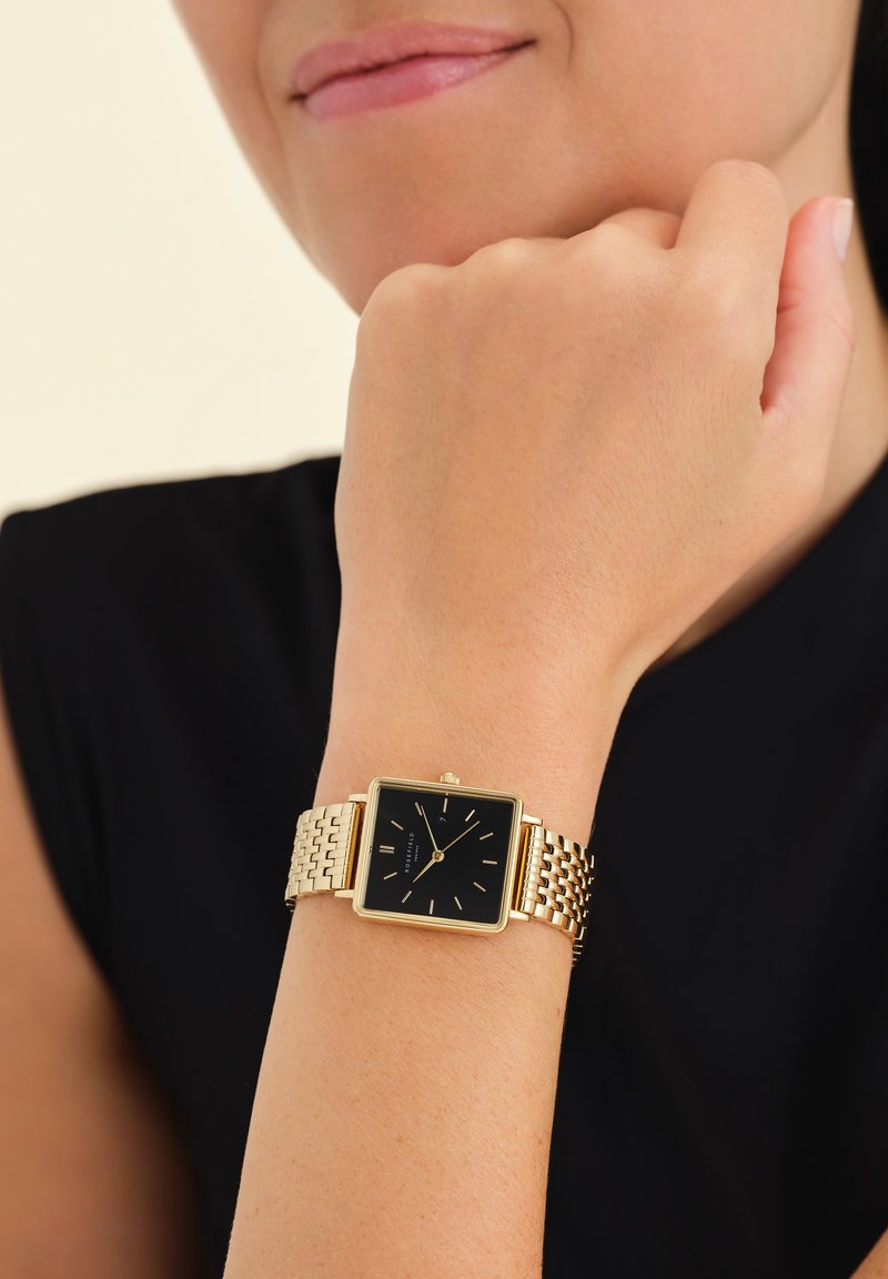 Goldfarbene Metallarmbanduhr mit schwarzem quadratischem Ziffernblatt, minimalistischen Stundenmarkierungen und einem geflochtenen Armband.