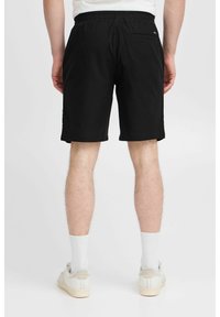 Shorts noirs à taille élastique avec poches latérales et une poche arrière, fabriqués en tissu doux, présentés de dos. Portés avec des chaussettes blanches et des chaussures.