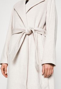 Abrigo de mezcla de lana beige claro con cuello de chal, cintura ceñida con cinturón y bolsillos laterales; textura suave y corte relajado.
