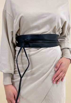 Black leather belt with a wide strap and thin tie detail, worn over a beige draped top, smooth texture, minimalistic design.