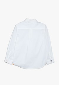 Valkoinen pitkähihaiset paita, takaapäin näkymä. Ominaisuuksiin kuuluu perinteinen kaulus, napitettu hihansuu ja poimutus keskellä. Valmistettu kevyestä kankaasta.