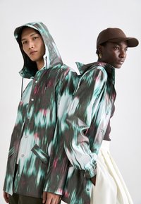 Multicolored waterproof jackets with an abstract pattern, featuring a hood, buttons, and pockets, worn by two models.