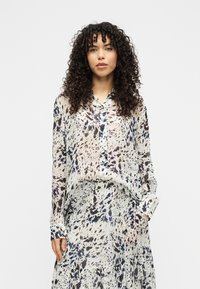 Αέρινη, διάφανη μπλούζα και φούστα σε εκρού χρώμα με αφηρημένα μοτίβα στίλβωσης σε μπλε ναυτικό και καφέ. Μακριά μανίκια, μυτερός γιακάς, εμπρόσθιο κούμπωμα με κουμπιά.