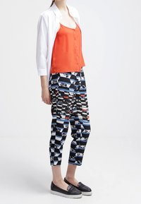 Camisole en satin orange avec des boutons, superposé sous une chemise blanche. Pantalons cropped noir et blanc à motifs avec des accents rouges. Chaussures noires à enfiler.