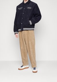 Uomo che indossa un giubbotto in corduroy blu navy con testo bianco, pantaloni larghi in corduroy beige e sneakers bianche con suole in gomma e lacci blu navy.