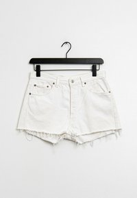 Pantalones cortos de mezclilla blanca con dobladillo deshilachado, diseño de cinco bolsillos, herrajes metálicos y cierre con botón, colgando de una percha negra.
