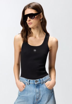 Donna che indossa una canotta nera con un piccolo emblema, jeans blu, occhiali da sole neri oversize e orecchini a cerchio dorati, con le mani nelle tasche.