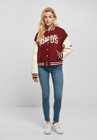 Burgundy varsity jacket with cream sleeves, featuring white lettering and striped ribbing. Worn over blue skinny jeans and white sneakers.