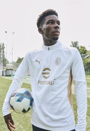 Chaqueta deportiva blanca con detalles dorados, cuello con cremallera y logo. Balón de fútbol negro y azul en la mano, con fondo de hierba verde.