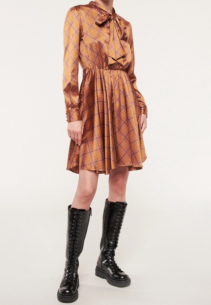 Kvinna i en brun och orange rutig klänning med en framåtbåge, kombinerad med knähöga svarta snörstövlar, stående på en vit bakgrund.