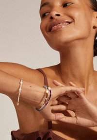 Vier Perlenarmbänder in verschiedenen Farben und Materialien schmücken ein Handgelenk, ausgestattet mit Perlen, bunten Steinen und komplexen Designs auf einem neutralen Hintergrund.